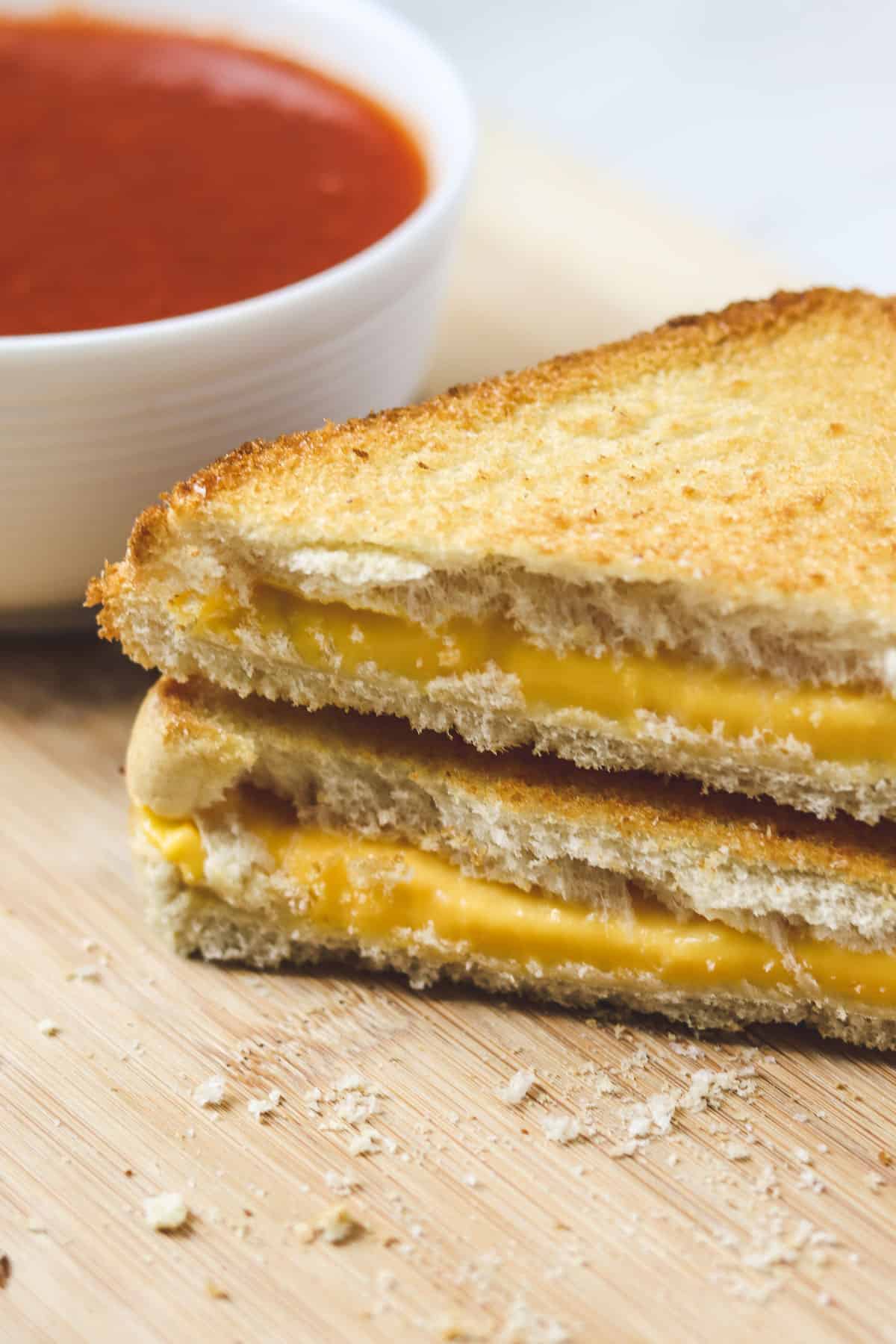 grilled cheese cut in half and stacked on a wood cutting board with tomato soup in the background