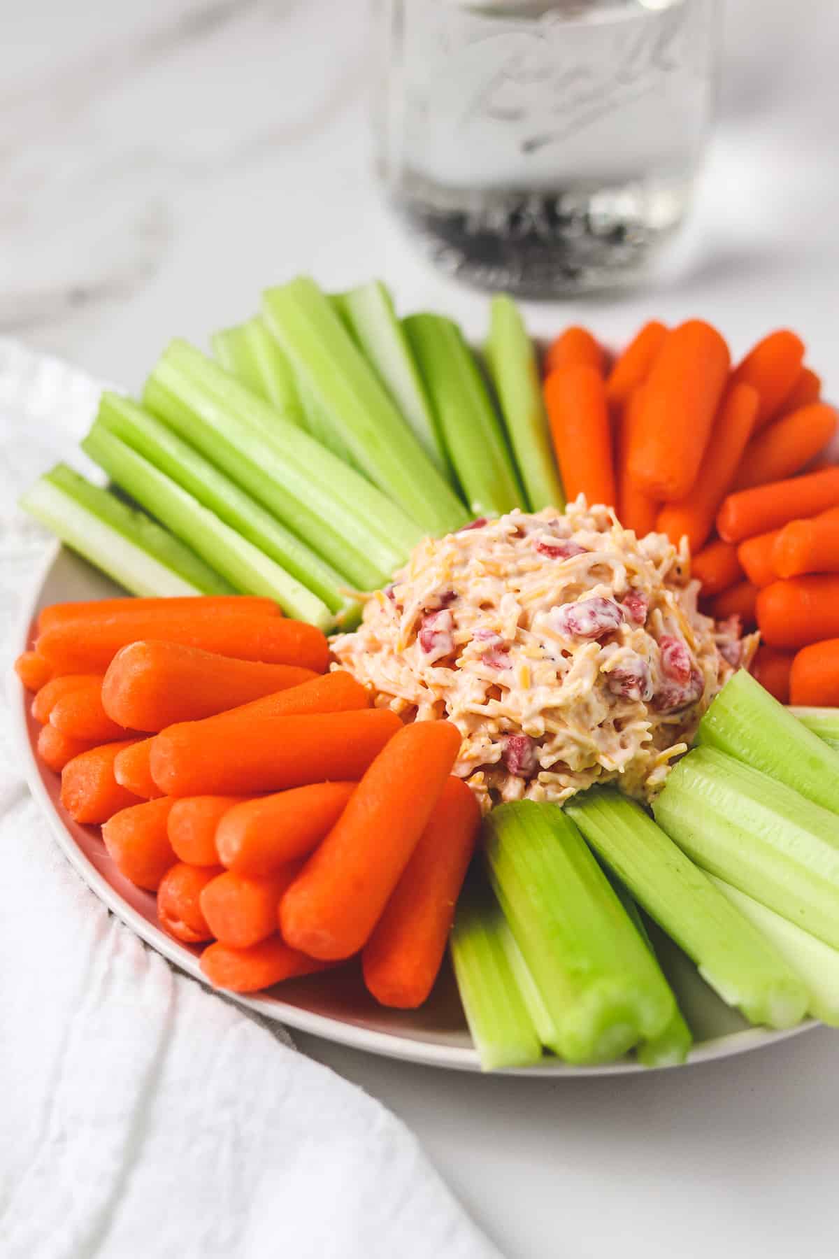 pimento cheese on a plate with carrots and celery with white napkin and water in mason jar