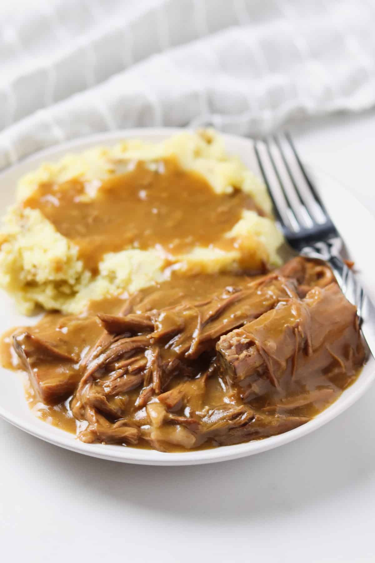 pot roast with gravy on a white plate with mashed potatoes and a fork