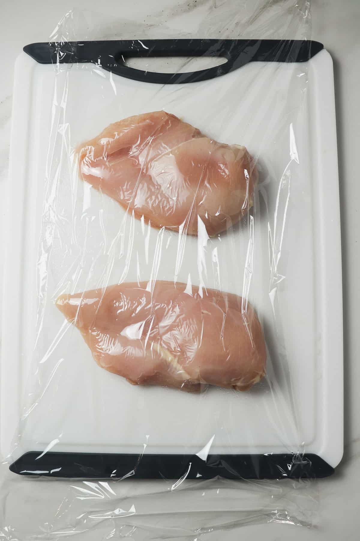 pounding chicken breasts thin under a sheet of plastic wrap on a white cutting board