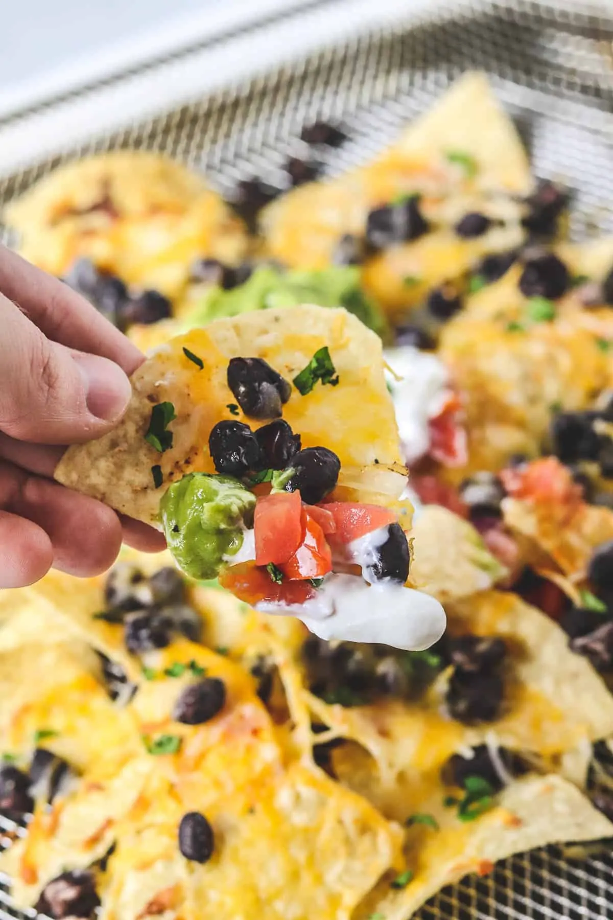 holding a chip of nachos with black beans, cheese, guacamole, sour cream, and pico de gallo